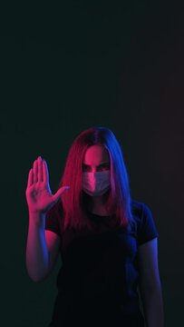 Stop sign. Keep distance. Woman in protective mask neon light glow showing forbidden hand gesture looking at camera isolated on black vertical. Quarantine measures. Pandemic restriction. New normal