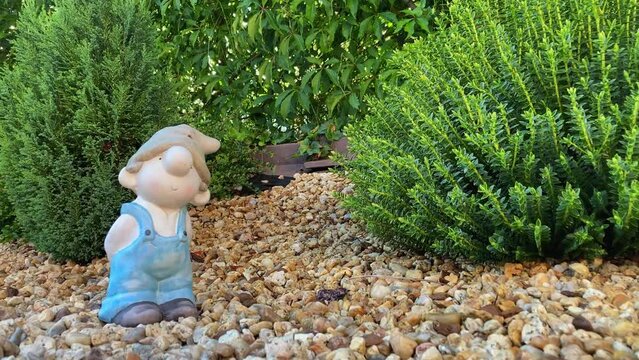 A cute garden gnome in work overalls standing in a flower bed, decorative stones, plants.