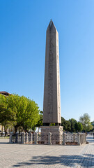 Obelisk of Theodosius seen from one end of the square, with a completely blue sky in the morning. With trees around.