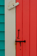 A vibrant teal green house with cream color trim and a vibrant orange shutter door. The closed door has a hook and latch attached. The locks are rusty vintage iron. The wall is narrow clapboard boards