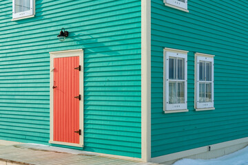 The exterior of a vibrant orange color single traditional wooden shutter door with cream color trim on a teal green clapboard wall of a historic house with two single hung windows with lace curtains. - obrazy, fototapety, plakaty