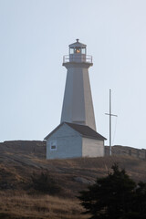 The top of a white colored vintage round lighthouse, lantern room, and tower containing storm glass panes. There's a mound of dried grass in the foreground. The background is a blue and cloudy sky.