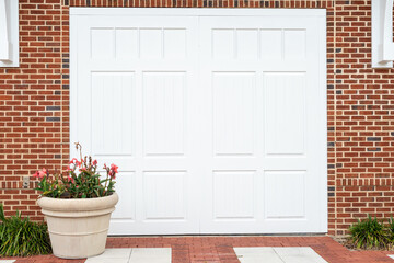 A white metal garage panel door in a red brick house. There's a large clay pot of flowers in from of the door. The entrance to the building has bricks on the ground with two rows of white blocks.  
