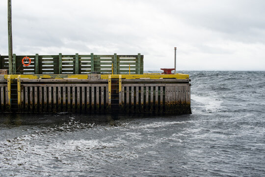A wooden dock with yellow trim. The dock decking is made of pressure treated wood.  The strong ocean waves are beating against the marina. There's a  red bollard, life ring, and pole on the marina. 