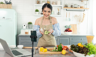 Asian woman food blogger cooking salad in front of smartphone camera while recording vlog video and live streaming at home in kitchen.