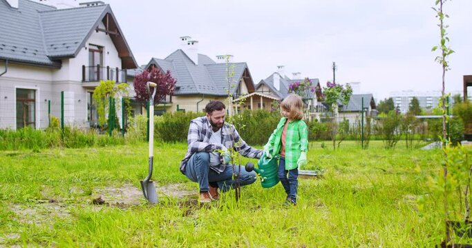 Caucasian father and cute little son watering planted tree with pot and giving high five to each other at garden. Summer work in orchard with kid. Summerhouse yard. Planting trees.