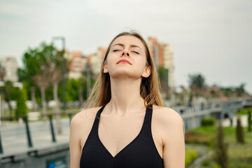 Cute brunette woman wearing sports bra standing on city park, outdoors relaxing with eyes closed, feeling alive, breathing, dreaming.