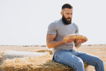 Happy bearded man farmer in jeans holding fresh fragrant bread and sitting top on the bale around...