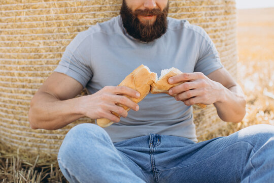 Happy bearded man farmer in jeans holding fresh fragrant bread and sitting near the bale around sunny sun field.