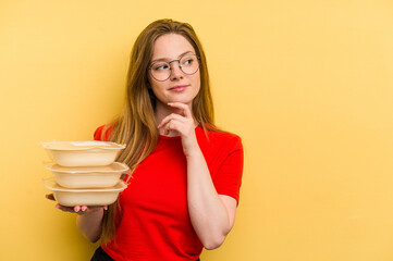 Young caucasian woman holding tupper isolated on yellow background looking sideways with doubtful and skeptical expression.