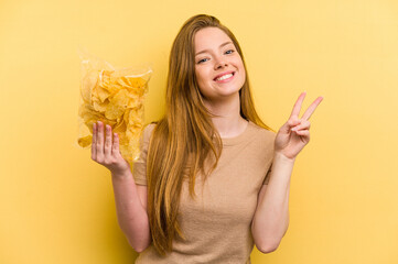 Young caucasian woman holding a bag of chips isolated on yellow background joyful and carefree...