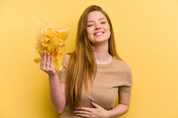 Young caucasian woman holding a bag of chips isolated on yellow background laughing and having fun.