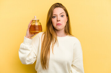 Young caucasian woman holding a honey isolated on yellow background shrugs shoulders and open eyes...