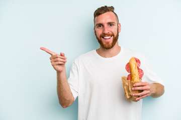 Young caucasian man eating a sandwich isolated on blue background smiling and pointing aside, showing something at blank space.