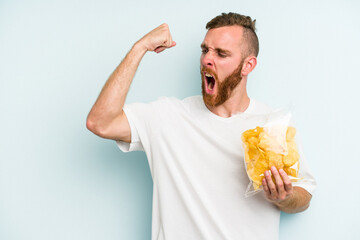 Fototapeta premium Young caucasian man holding crisps isolated on blue background raising fist after a victory, winner concept.