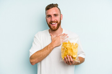 Young caucasian man holding crisps isolated on blue background laughs out loudly keeping hand on chest.