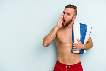 Young caucasian man going to the beach holding a towel isolated on blue background is saying a secret hot braking news and looking aside