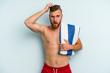 Young caucasian man going to the beach holding a towel isolated on blue background being shocked,...