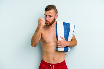 Young caucasian man going to the beach holding a towel isolated on blue background showing fist to camera, aggressive facial expression.