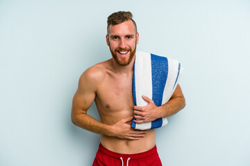 Young caucasian man going to the beach holding a towel isolated on blue background laughing and having fun.