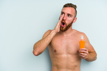 Fototapeta premium Young caucasian man holding solar cream isolated on blue background is saying a secret hot braking news and looking aside