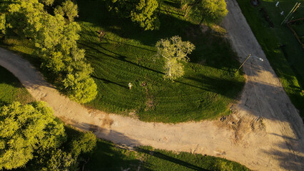 Aerial top down shot of tractor cutting grass in park during sunny day in the evening - Idyllic landscape with green trees in summer and sandy path
