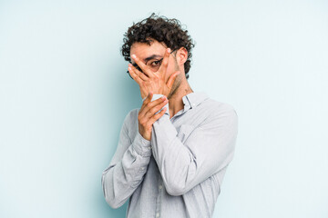 Young caucasian man isolated on blue background blink through fingers frightened and nervous.