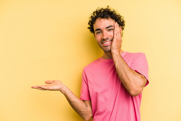 Young caucasian man isolated on yellow background holds copy space on a palm, keep hand over cheek. Amazed and delighted.