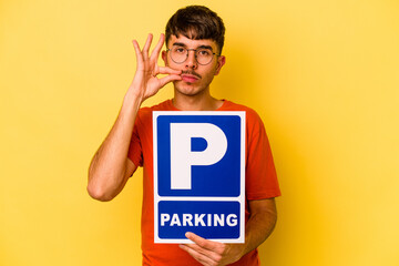 Young hispanic man holding parking placard isolated on yellow background with fingers on lips...