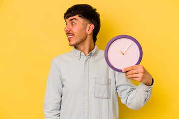 Young hispanic man holding a clock isolated on yellow background looks aside smiling, cheerful and pleasant.
