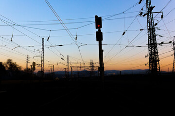 Red signal semaphore on the railroad on a dark background at dusk. Black silhouettes catenary and masts against the background of the sunset sky