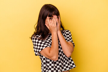 Young caucasian woman isolated on yellow background blink at the camera through fingers, embarrassed covering face.