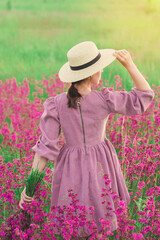 A girl in a straw hat and a purple dress walks through a field with blooming purple flowers at sunset