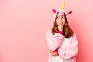 Little caucasian girl wearing a unicorn pajama isolated on pink background looking sideways with doubtful and skeptical expression.