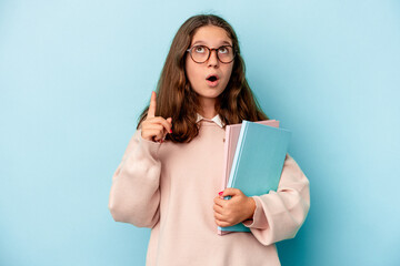 Little caucasian student girl holding books isolated on blue background pointing upside with opened mouth.