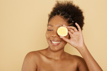 Beautiful afro american female model holding half a lemon in front of her face on a beige background. The concept of caring cosmetics with vitamin c, skin care and spa.