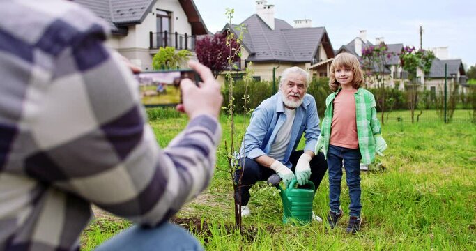 View over shoulder on man taking photo with smartphone of grandfather and grandson posing in garden after planing trees. Male making picture with phone camera of father and small son.