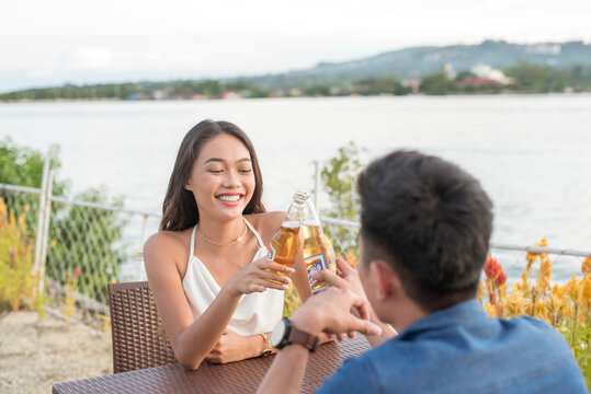 Two young attractive people make a toast with their beer bottles while at a an outdoor bar with waterfront views. A first date going very well, excellent chemistry.