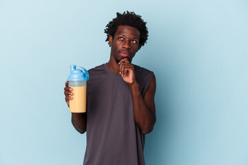 Young african american man holding protein milkshake isolated on blue background looking sideways...