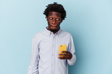 Young african american man holding mobile phone isolated on blue background confused, feels...