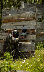 game of paintball, a male player sits near a wooden fence and shoots colored balls. summer in the grass sunny day. overall plan