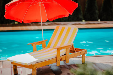 Stripped summer beach chair with red sun umbrella against blue swimming pool.