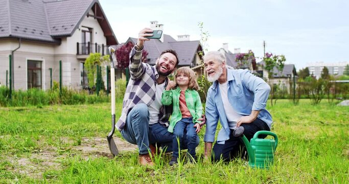 Cheerful man taking selfie photo with smartphone of grandfather, father, son and grandson sitting in garden and smiling to camera. Planting trees concept. Making picture of three generations.