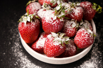 Strawberries in a plate with powdered sugar on a black background, close-up.