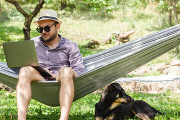 Man in hat and sunglasses sitting in hammock and using his laptop while his dog lying down under hammock. - Powered by Adobe