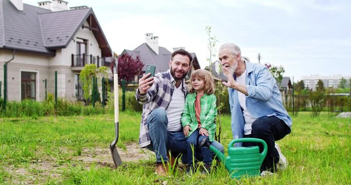 Caucasian kid with father and grandfather sitting in garden and videochatting via smartphone. Small boy showing planting trees at summerhouse. Videochat on phone. Call by video.