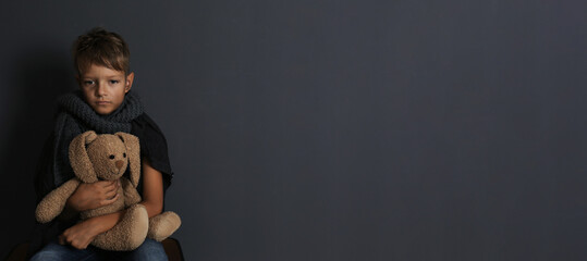 Poor boy sitting with toy rabbit on dark background, space for text. Banner design