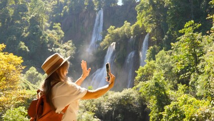 Happy carefree woman tourist talk with friend or parents via selfie video call on smartphone. Positive young female traveler in straw hat waving hand at camera standing near waterfall. Slow motion.