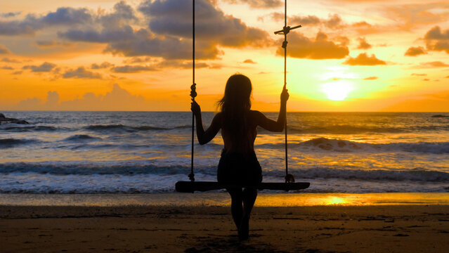 Young woman swinging sitting on wooden swing with ropes on sandy beach, sea sunset background. Girl silhouette enjoy outdoors freedom, summer. Travel, tourist, holidays, vacations concept. Back view.