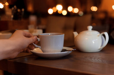White cup with teapot. The hand holding the cup of tea standing on saucer in soft focus on naturally blurred background. Coffee, tea house, bokeh lights. The concept of a cozy pastime